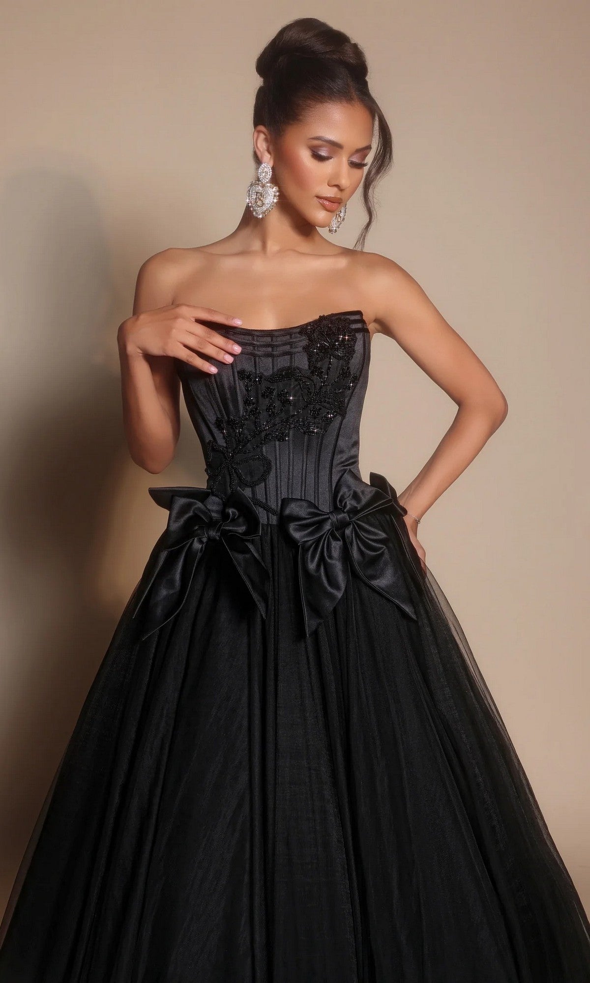 A woman poses against a neutral background wearing the Portia and Scarlett PS26102 strapless ball gown with a corset bodice, black floral appliqué, satin bows at the waist, and statement earrings.